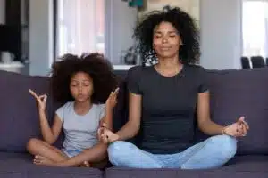 A Black Mother and Daughter Meditate on the couch during a Mental Health Day