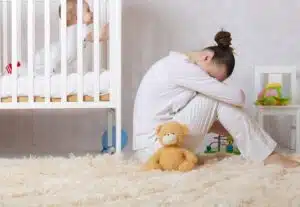 A young mother sits on floor near crib with baby, suffering from postpartum depression