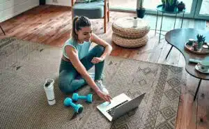 A sporty woman in sportswear is sitting on the floor with dumbbells and a protein shake or a bottle of water and is using a laptop at home in the living room.