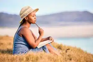 Young black woman wearing a hat thinking while writing in a journal overlooking the shore