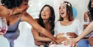 Excited young female friends touching pregnant woman's belly at baby shower party. Laughing pregnant woman sitting with friends touching her tummy.