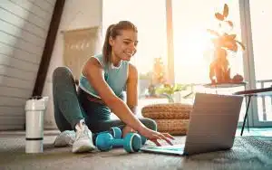 Young woman in workout gear sitting on floor and looking at laptop for workout inspiration