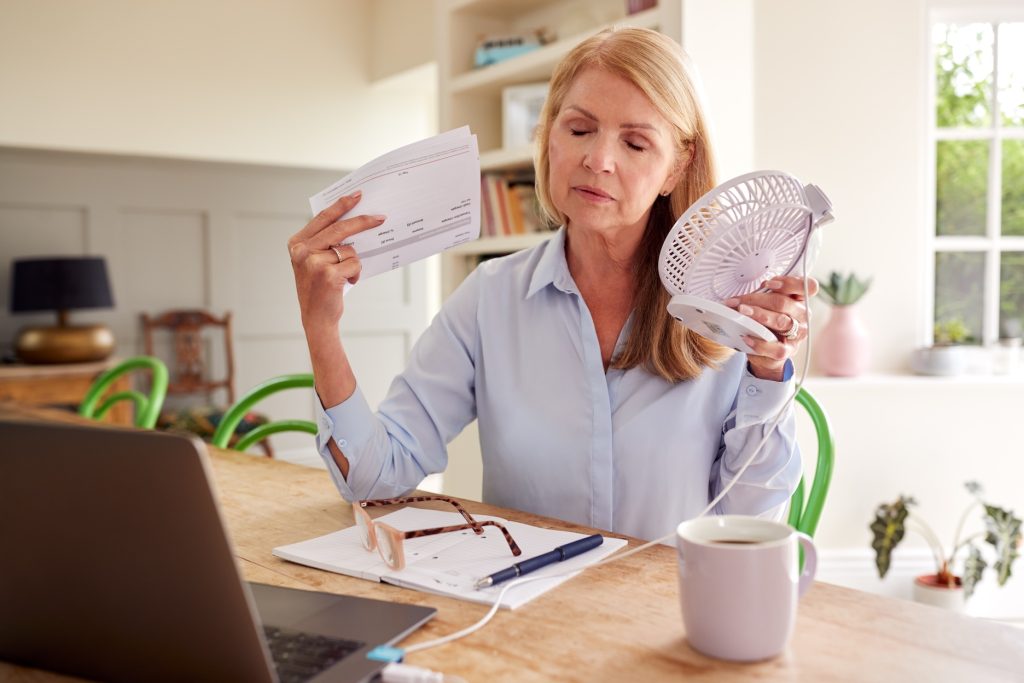 A woman in her kitchen in front of her laptop having a hot flash with a fan in one hand and bills in the other.