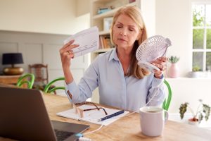 A woman in her kitchen in front of her laptop having a hot flash with a fan in one hand and bills in the other.