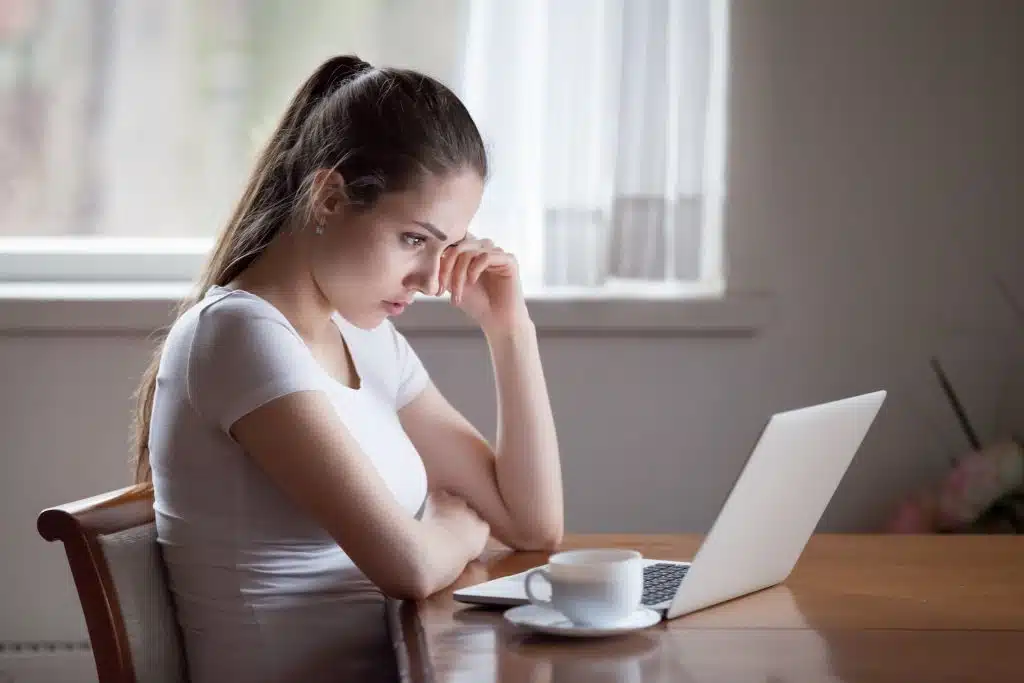 A thoughtful, young woman with brown hair in a ponytail in front of a laptop and cup of coffee...meant to convey online therapy