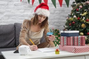 A woman calculates holiday budget with a Christmas tree and presents beside her
