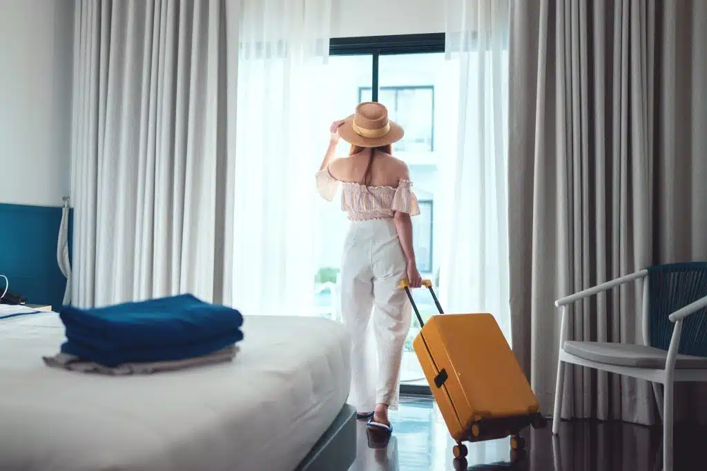 A woman wearing a hat with rolling luggage looks out the window of her guest room.
