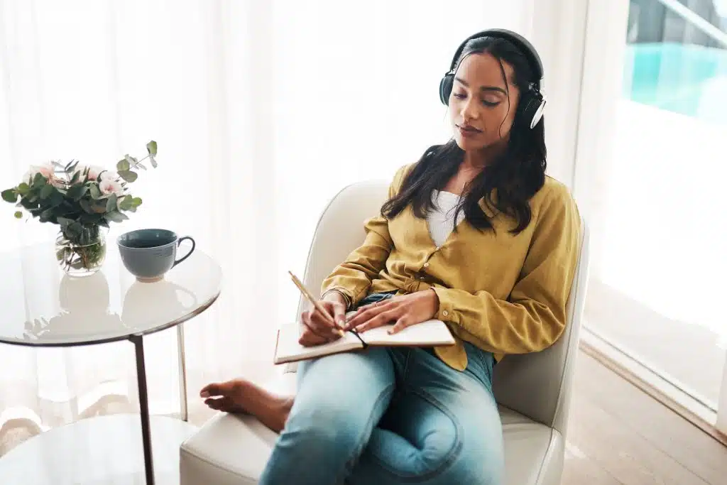 A young woman wearing headphones sits in a chair while writing in a journal.