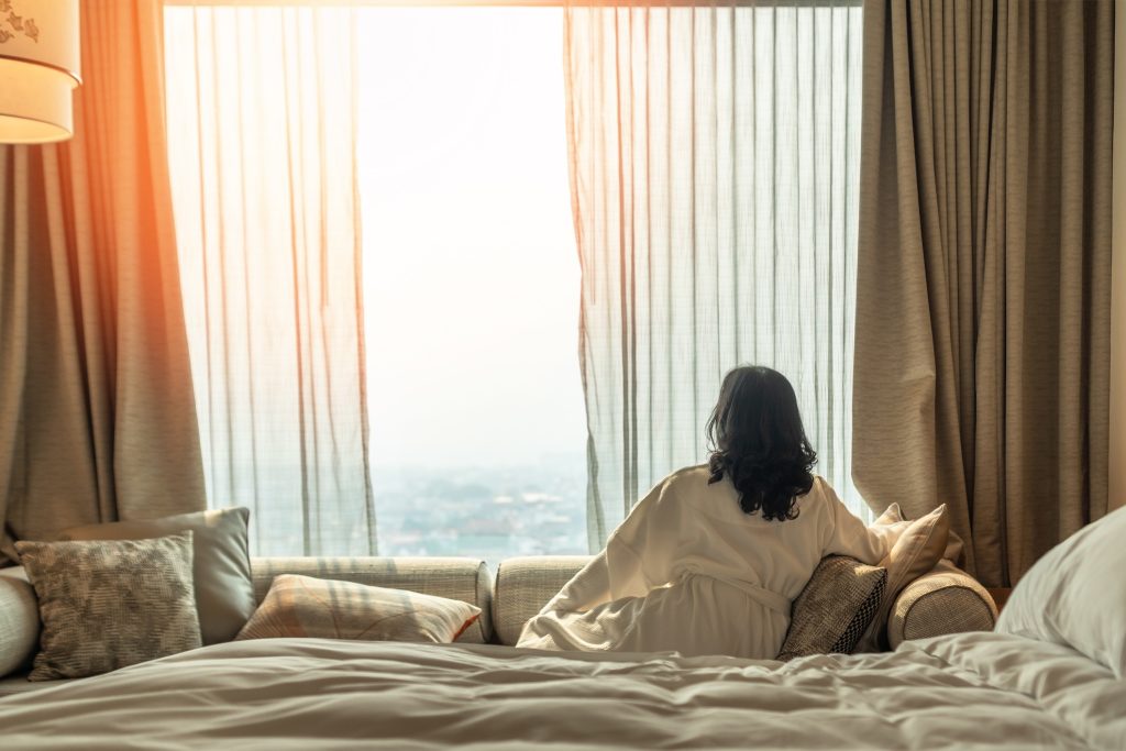 A woman relaxing in her hotel room looking out the window as the sun rises.